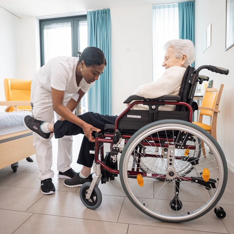 Assistentin Gesundheit und Soziales hilft einer Seniorin in den Rollstuhl